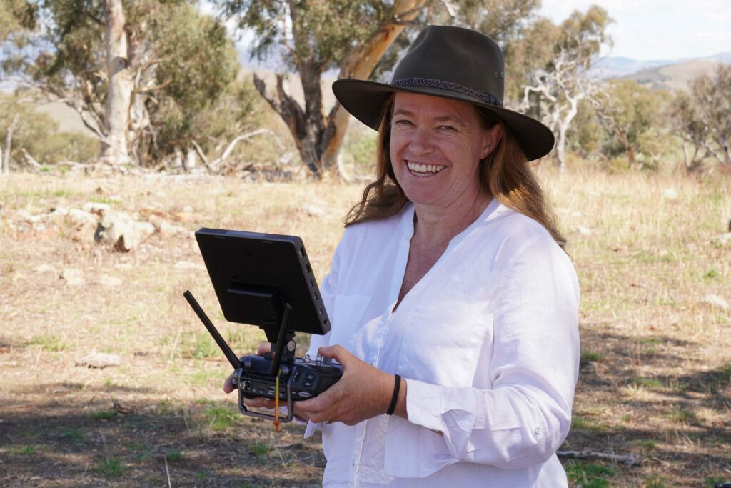 debbie, the founder of wildlife drones is pictured here. she wears a white blouse and broad brim hat and is smiling at the camera about to fly a drone